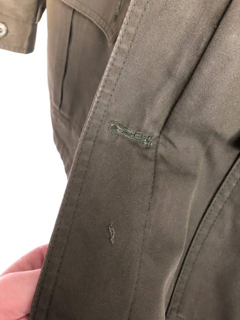 May include: Close-up of a dark green jacket with a button closure. The button is made of metal and has a loop design.