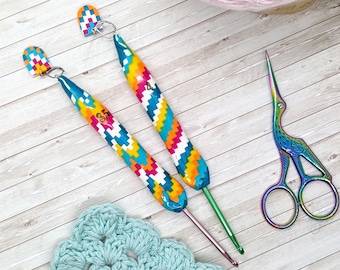May include: Two crochet hooks with vibrant, patterned handles and metal hooks. One hook is marked with the number 3.5. A pair of iridescent scissors and a light blue crocheted doily are also visible, suggesting a crafting theme.