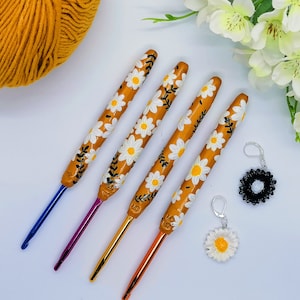 May include: Four crochet hooks with daisy flower designs painted on a yellow background. The hooks are different colors: blue, purple, gold, and orange. Two daisy flower charms are also pictured.