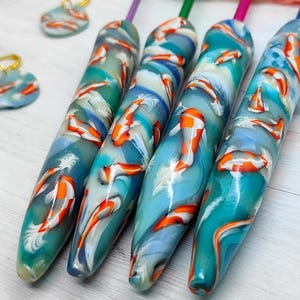 May include: Four crochet hook handles with a koi fish design in shades of blue, white, and orange. The handles have different colored tops. Two matching stitch markers are also visible.