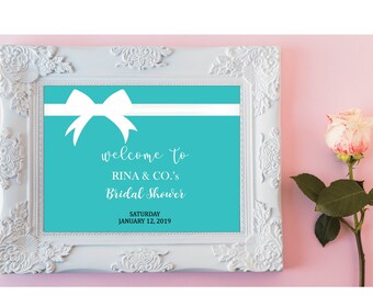 May include: A turquoise blue welcome sign with a white ribbon and bow. The text reads "welcome to RINA & CO.'s Bridal Shower, Saturday January 12, 2019".