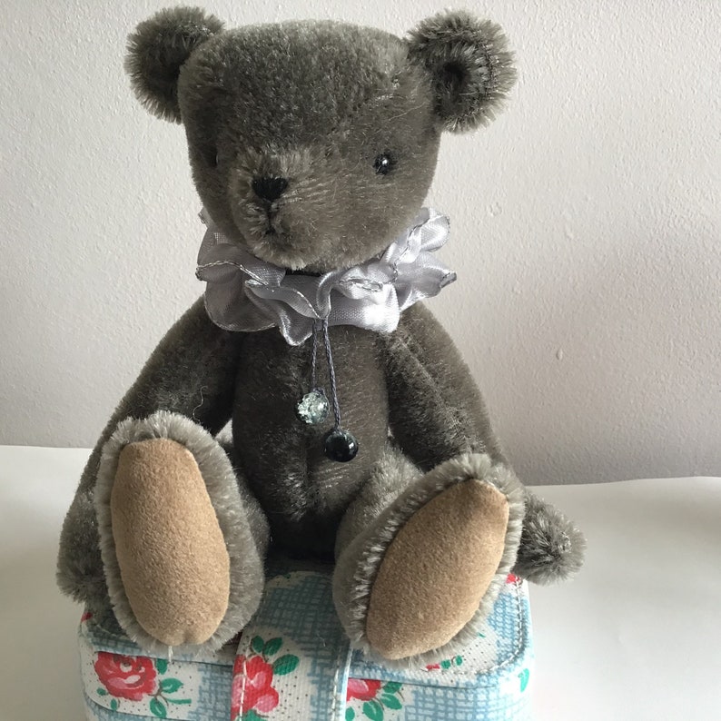 May include: A gray teddy bear with a silver satin bow around its neck. The bear is sitting on a blue and white checkered fabric with floral accents.