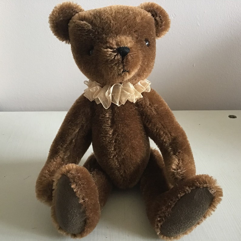 May include: A brown teddy bear with a beige lace collar. The bear is sitting with its paws in front of it.