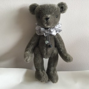 May include: A gray teddy bear with a silver ruffled collar and black bead accents. The bear is standing on a white surface.