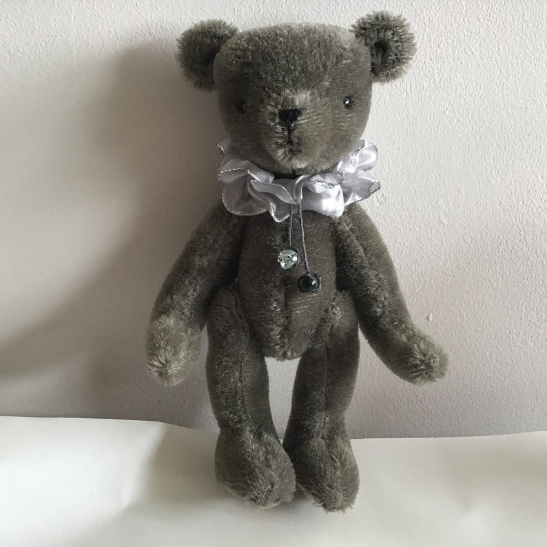 May include: A gray teddy bear with a silver ruffled collar and black bead accents. The bear is standing on a white surface.
