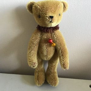 May include: A small, brown teddy bear with a brown and red ribbon around its neck. The bear has a small bell attached to the ribbon.