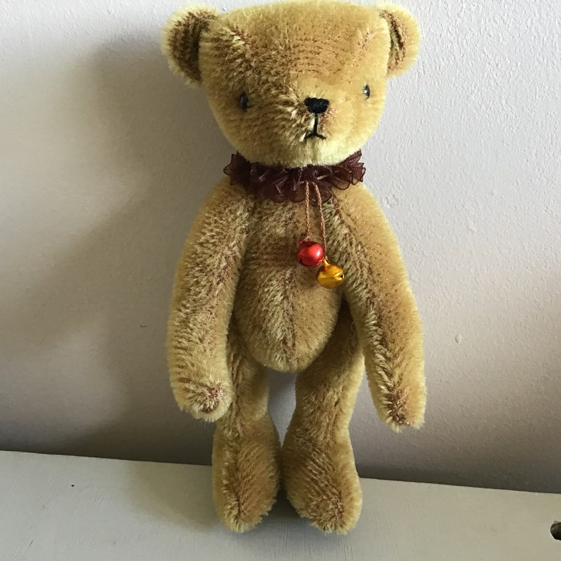 May include: A small, brown teddy bear with a brown and red ribbon around its neck. The bear has a small bell attached to the ribbon.