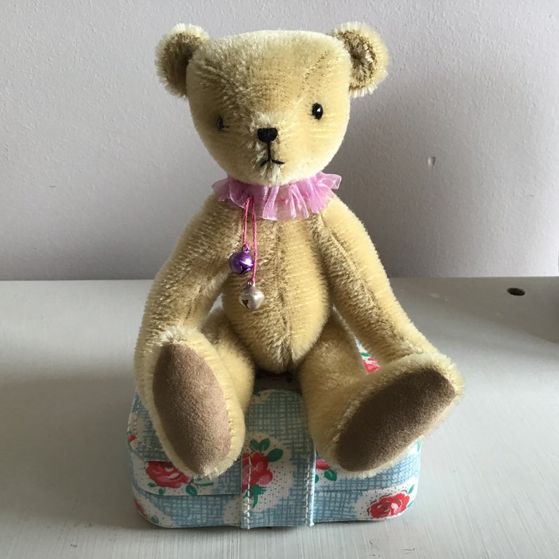 May include: A plush, beige teddy bear with a pink and purple ribbon around its neck. The bear is sitting on a blue and white floral patterned box.