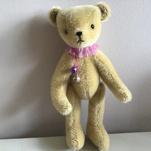 May include: A light brown teddy bear with a pink ruffled collar and a string of bells hanging from it.