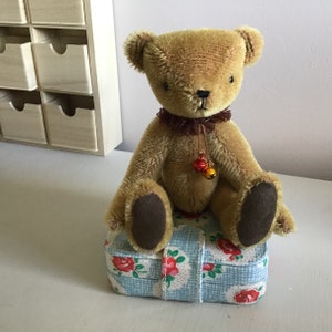 May include: A brown teddy bear with a brown velvet nose and brown eyes sits on a blue and white floral patterned box. The bear has a brown velvet bow around its neck with two small bells hanging from it.