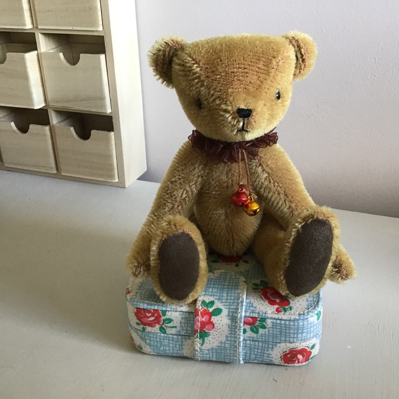 May include: A brown teddy bear with a brown velvet nose and brown eyes sits on a blue and white floral patterned box. The bear has a brown velvet bow around its neck with two small bells hanging from it.