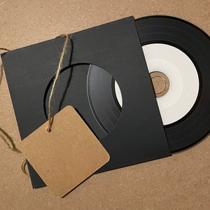 May include: A black square paper sleeve with a circular cutout revealing a black and white compact disc. A blank, square, light brown cardboard tag is attached to the sleeve with a thin piece of natural jute twine, hanging off the left side.