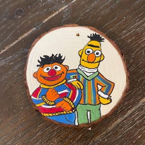 May include: A hand-painted wooden ornament featuring Sesame Street characters Big Bird and Elmo. Big Bird is wearing a yellow shirt with orange and green stripes and Elmo is wearing a red and blue striped shirt with a yellow collar.