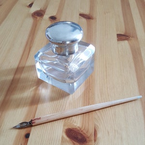 Può includere: Un calamaio di vetro trasparente con un coperchio d'argento su una superficie di legno. Una penna a immersione in legno è sdraiata accanto al calamaio.