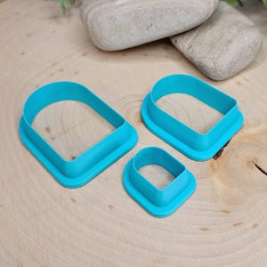 May include: Three turquoise cookie cutters in varying sizes, each with a rounded rectangular form. They are arranged on a light-coloured wooden surface, with two grey stones in the background.