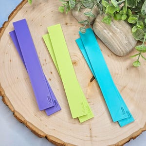 May include: Three colorful, textured strips in purple, lime green, and turquoise, each with a different width measurement in millimeters. The strips are arranged on a wooden surface, with greenery in the background.