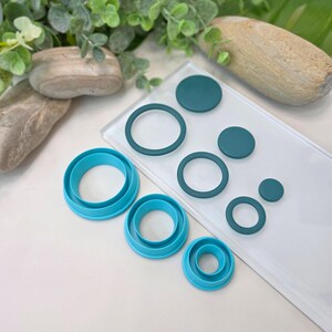 May include: A selection of teal and blue circle-shaped cookie cutters and teal polymer clay circles and rings. The cutters are nested in graduated sizes. The clay pieces are in a clear plastic sleeve. The background includes greenery and a light-coloured surface.