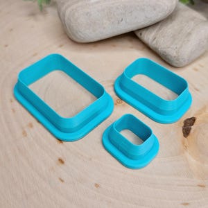 May include: Three turquoise rectangular cookie cutters of varying sizes are arranged on a light wood surface. The cutters have a raised, layered design. The largest cutter is at the top left, with the smallest at the bottom.