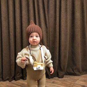 以下が含まれることがあります： 尖ったトップのついた茶色のニット帽とベージュのセーターとレギンスを着用した幼い子供。子供は首にストラップをつけたおもちゃのカメラを持っています。