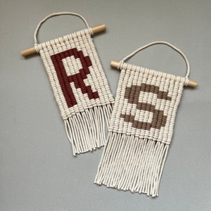 Könnte beinhalten: Zwei Makramee-Wandbehänge mit Holzleisten oben. Die Behänge sind aus naturfarbenem Garn gefertigt und zeigen die Buchstaben "R" und "S" in braunem und rotem Garn.