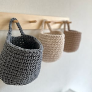 Pu&ograve; includere: Tre cesti in crochet appesi in grigio, beige e marrone. I cesti sono appesi a un rack di legno con ganci in ottone.