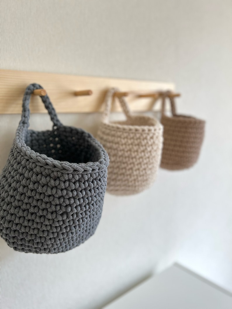 Pu&ograve; includere: Tre cesti in crochet appesi in grigio, beige e marrone. I cesti sono appesi a un rack di legno con ganci in ottone.