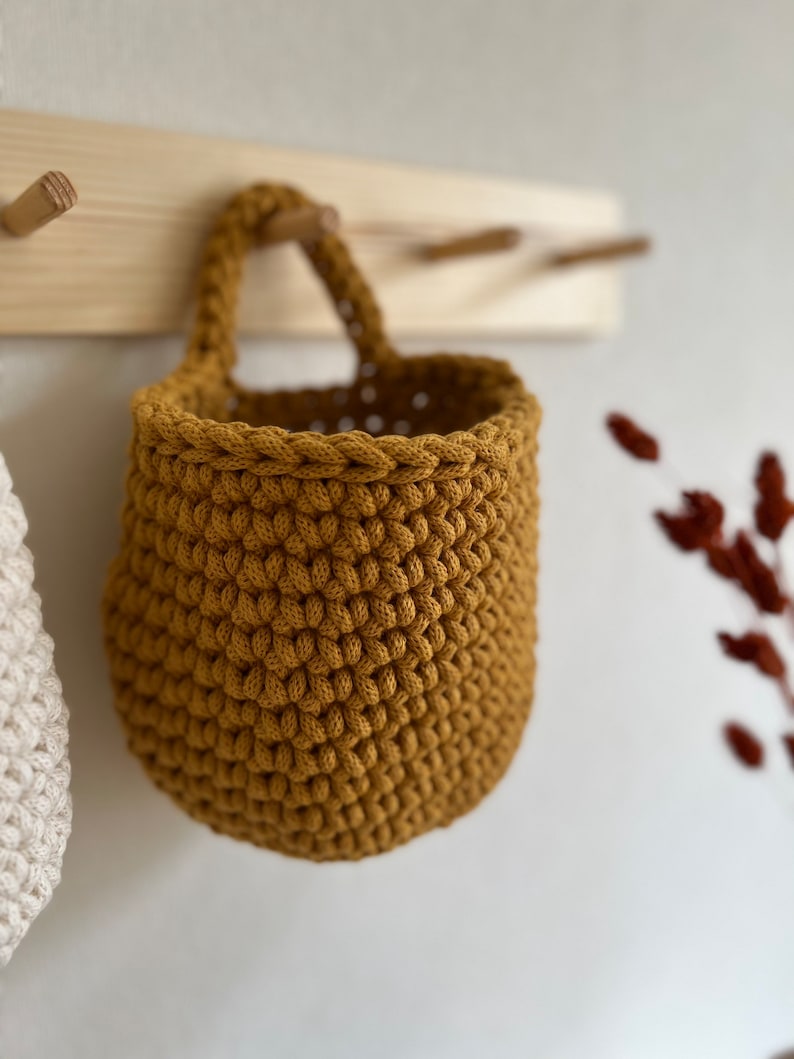 Pu&ograve; includere: Un cesto appeso in crochet di colore giallo senape con un manico in corda.
