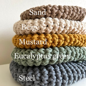 Pu&ograve; includere: Cinque sottobicchieri rotondi all'uncinetto in diversi colori, impilati uno sopra l'altro. I colori sono sabbia, beige, senape, verde eucalipto e acciaio.