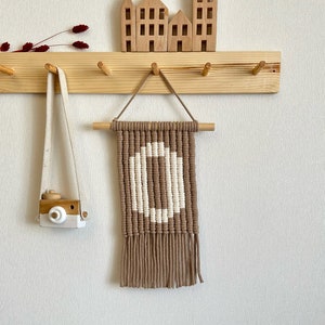 May include: A wooden coat rack with pegs, featuring a macrame wall hanging with a white pattern on a brown backdrop. A wooden toy camera hangs to the left. Wooden house ornaments sit on a shelf above.
