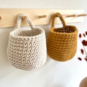 Pu&ograve; includere: Due cesti appesi all'uncinetto, uno beige e uno giallo senape, sono esposti su un portaoggetti da parete in legno.