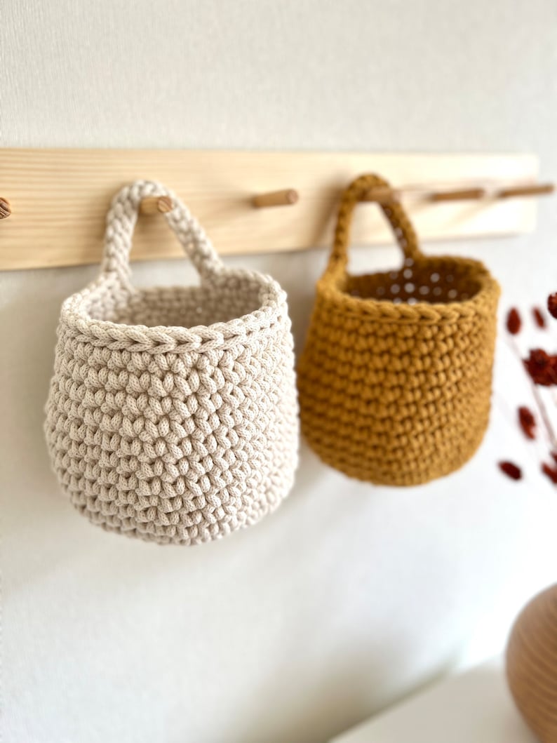 Pu&ograve; includere: Due cesti appesi all'uncinetto, uno beige e uno giallo senape, sono esposti su un portaoggetti da parete in legno.