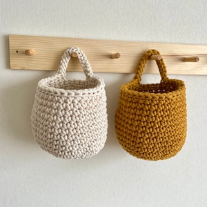 Pu&ograve; includere: Due cesti appesi all'uncinetto, uno beige e uno giallo senape, sono appesi a un rack di legno con ganci.