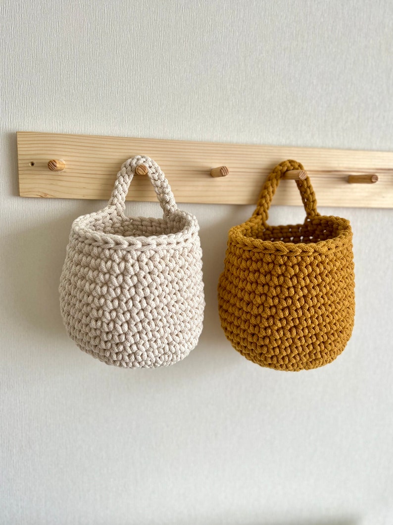 Pu&ograve; includere: Due cesti appesi all'uncinetto, uno beige e uno giallo senape, sono appesi a un rack di legno con ganci.