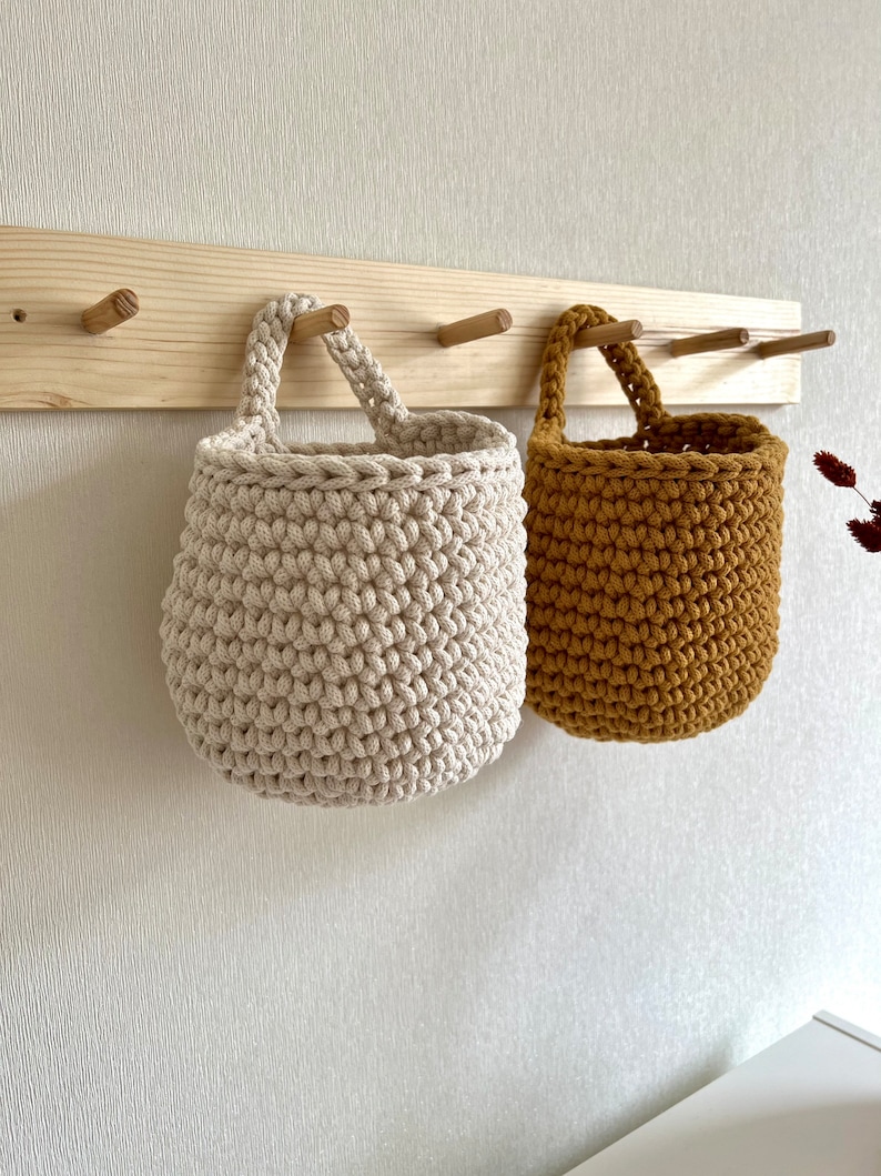 Pu&ograve; includere: Due cesti appesi all'uncinetto, uno beige e uno giallo senape, sono appesi a un portaoggetti da parete in legno con ganci.