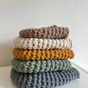 Pu&ograve; includere: Un mucchio di cinque cuscini rotondi all'uncinetto in vari colori, tra cui marrone, beige, giallo senape, verde e grigio.