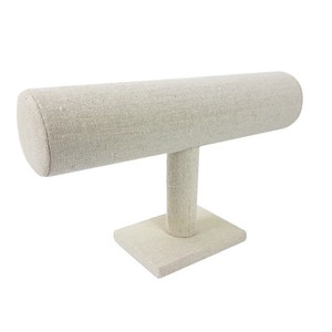 May include: A beige fabric-covered jewelry display stand. The stand features a horizontal, cylindrical bar supported by a vertical post and a square base. The neutral color and simple design make it suitable for showcasing bracelets or other accessories.