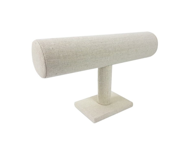 May include: A beige fabric-covered jewelry display stand. The stand features a horizontal, cylindrical bar supported by a vertical post and a square base. The neutral color and simple design make it suitable for showcasing bracelets or other accessories.