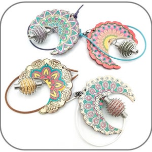 Può includere: Tre paia di orecchini a forma di mezzaluna con disegni di mandala. Ogni orecchino ha una gabbia di metallo con un perno colorato all'interno. Gli orecchini sono su uno sfondo bianco.
