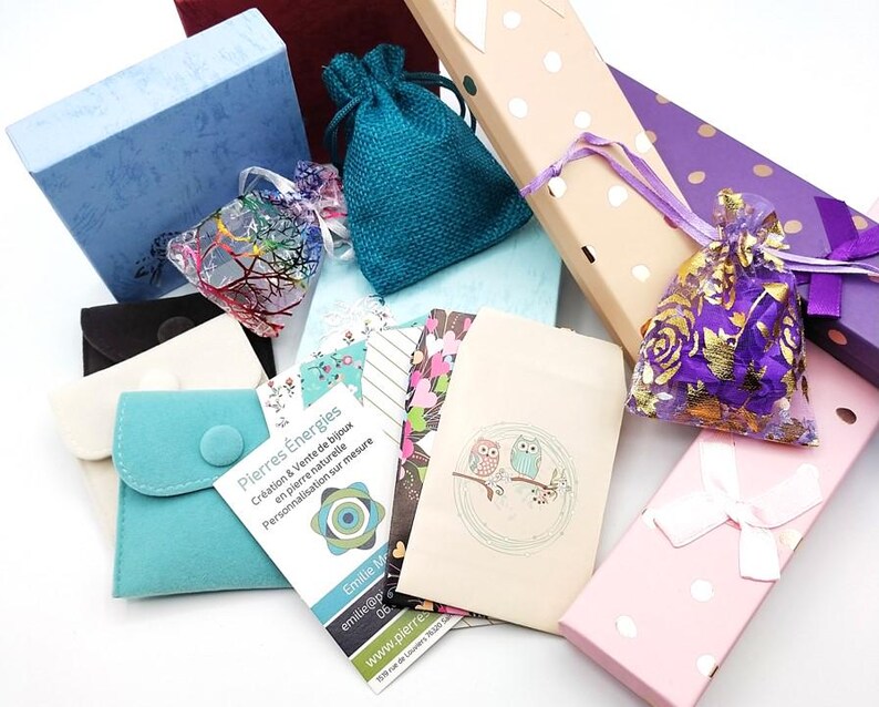 K&ouml;nnte beinhalten: Eine Auswahl an kleinen Geschenkboxen und Beuteln in verschiedenen Farben und Mustern, darunter Blau, Lila, Rosa und Braun. Einige Boxen haben Punkte, w&auml;hrend andere Blumenmuster haben. Die Boxen sind mit kleinen Gegenst&auml;nden gef&uuml;llt, wie z. B. Schmuck, Kristallen und anderen Kleinigkeiten. Der Text auf einer der Karten lautet "Pierres Energies Creation & Vente de bijoux en pierre naturelle Personnalisation sur mesure Emille M emiliep 06 www.pierre"