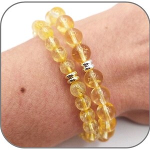 Può includere: Due braccialetti in pietra gialla con accenti argentati. I braccialetti sono fatti di perline rotonde e sono impilati sul polso di una persona.