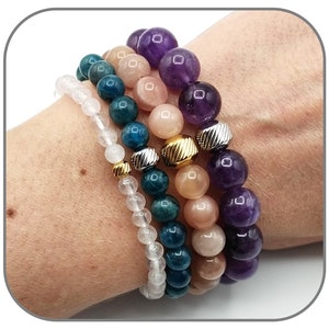 May include: Four stacked bracelets with different colored beads. The top bracelet has purple amethyst beads, the second bracelet has peach sunstone beads, the third bracelet has teal apatite beads, and the bottom bracelet has white quartz beads.