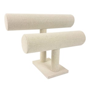 May include: A beige fabric jewelry display stand with two horizontal bars. The stand has a rectangular base and two vertical supports. The bars are cylindrical and designed to hold bracelets or necklaces. The neutral color makes it suitable for various jewelry styles.