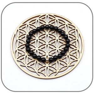 May include: Black beaded bracelet with a gold charm, displayed on a wooden circular background with a geometric pattern.