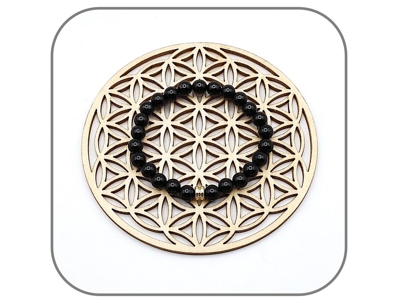 May include: Black beaded bracelet with a gold charm, displayed on a wooden circular background with a geometric pattern.
