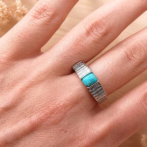 May include: A silver ring featuring a rectangular turquoise stone set in the center. The band has a textured, vertical grooved pattern across its surface, providing a distinct contrast to the smooth, bright blue surface of the inlaid stone.