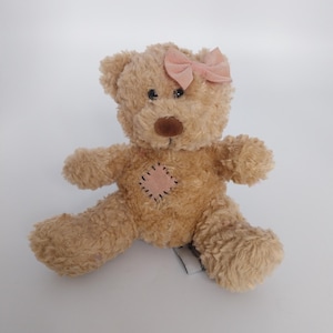 May include: A light brown teddy bear with a pink bow, brown nose, and black eyes. It has a stitched patch on its chest. The bear is sitting with its arms and legs spread. A classic children's toy.