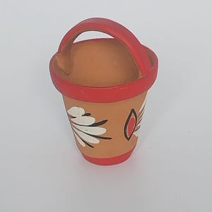 Puede incluir: Un pequeño cubo de cerámica de color terracota con borde y asa rojos. El cubo está decorado con diseños florales blancos y negros y una forma de lágrima roja. Es un artículo decorativo en miniatura.