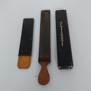 May include: Three vintage leather razor strops, one with a wooden handle and two in their original black boxes. The box on the right has the text "P.C.L. BUFFED AND EDGE STROP" printed on it.