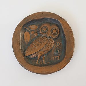 Puede incluir: Un plato redondo de cerámica de color cobre con una imagen en relieve de un búho con ojos grandes y una rama con bayas. El búho está mirando hacia la derecha y tiene un diseño detallado. El plato tiene un esmalte verde y las letras "AOE" son visibles en la parte inferior derecha.