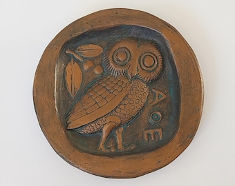 May include: A round, copper-colored ceramic plate with a raised image of an owl with large eyes and a branch with berries. The owl is facing right and has a detailed design. The plate has a green glaze and the letters "AOE" are visible on the bottom right side.